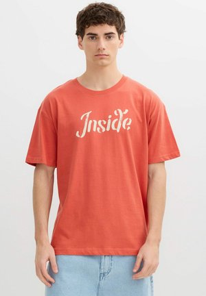 Joven con cabello castaño corto y rizado, vistiendo una camiseta naranja con la palabra "Inside" en letras blancas y vaqueros azul claro.