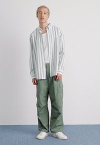 Herre-outfit bestående af en lysegrøn og hvidstribet langærmet skjorte, en hvid t-shirt underneath og cargo-lignende grønne bukser. Hvide sneakers.