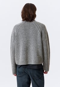 Une personne avec des cheveux bruns à mi-longueur porte un pull à rayures verticales gris et noir et un jean bleu foncé, de dos sur fond blanc.