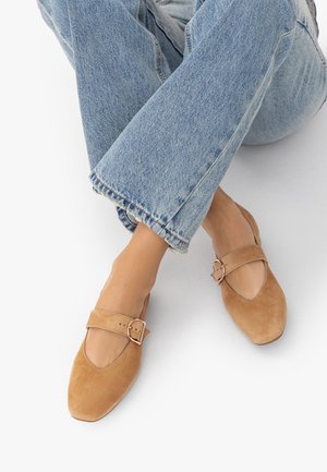 Gekreuzte Füße, die hellblaue Jeans aus Denim und beige Wildleder-Mary-Jane-Ballerinas mit goldener Schnalle auf weißem Boden tragen.