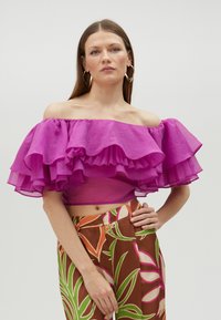 Fuchsia off-shoulder top med lagdelte flæser, parret med mønstrede brune bukser med farverige bladmotiver.