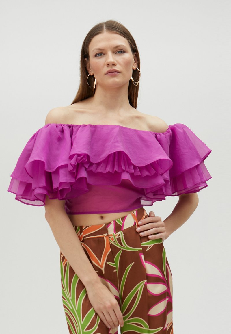 Fuchsia off-shoulder top med lagdelte flæser, parret med mønstrede brune bukser med farverige bladmotiver.