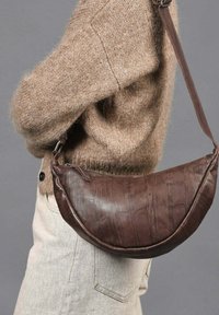 Bolso cruzado de cuero marrón con forma redondeada, acabado texturizado y correa desmontable, llevado sobre un suéter de punto beige.