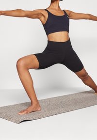 Czarny stanik sportowy i czarne szorty o wysokim stanie, noszone przez osobę w pozycji wojownika na teksturowanej macie do jogi z neutralnym tłem.