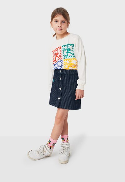 Langärmliges weißes Hemd mit bunten Tiermotiven, kombiniert mit einem dunkelblauen gestreiften Rock mit Knöpfen und grünen Akzenten. Weiße Sneakers und rosa Socken.