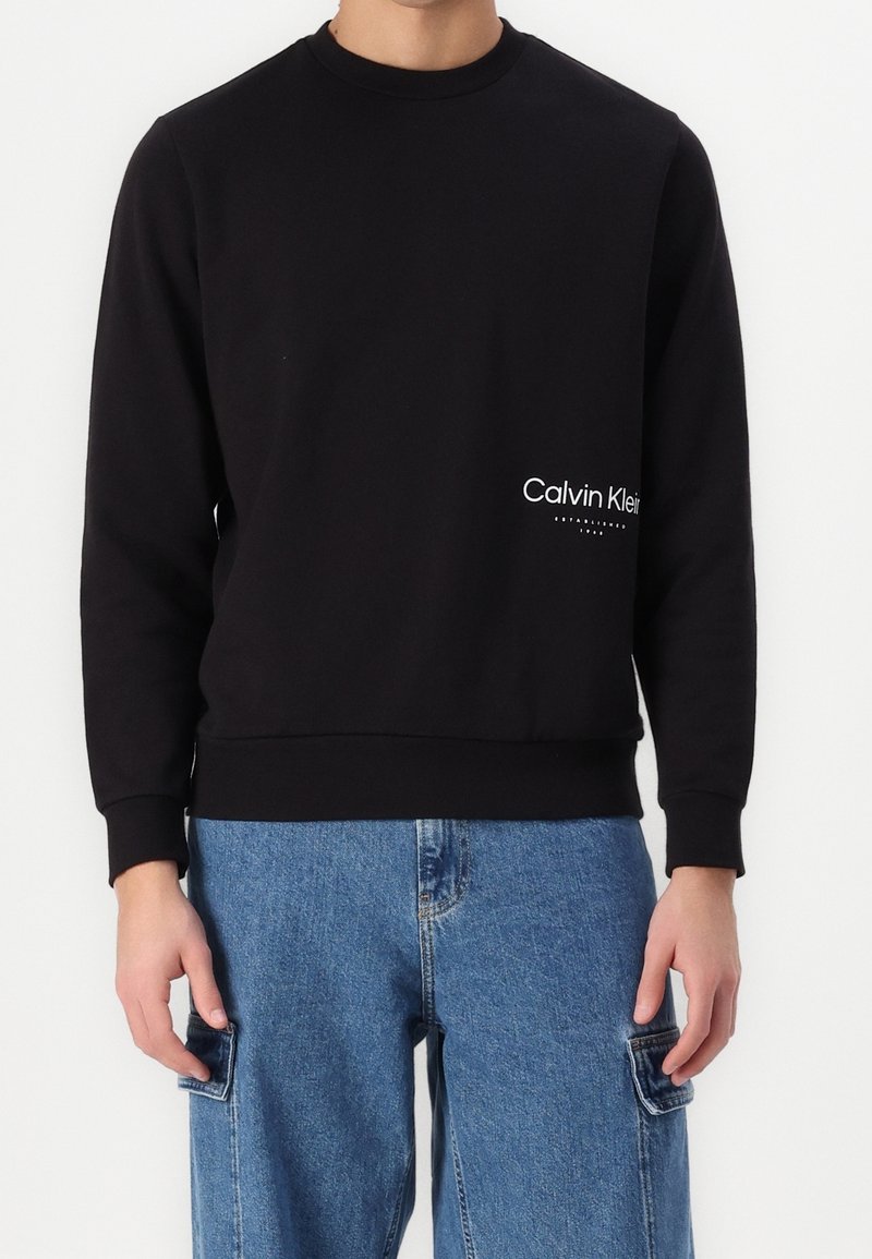 Persona que lleva un sudadera negra de Calvin Klein con el logo en la parte inferior derecha y unos pantalones vaqueros cargo azules con bolsillos laterales.