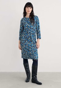 Rochie cu model floral în nuanțe de albastru, verde și bleumarin. Are mâneci trei sferturi și o croială lejeră, asortată cu cizme negre până la genunchi.