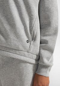 Felpa grigia con tessuto strutturato, polsini aderenti e un sottile dettaglio del logo vicino all'orlo. Il design include una cucitura curva.