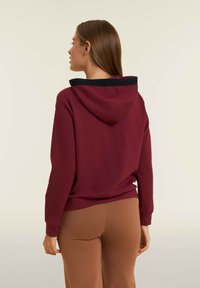 Sudadera burdeos con un cuello negro, que presenta un bolsillo canguro y puños de canalé. La tela parece suave y texturizada. Pantalones marrones visibles abajo.