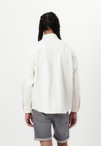 Camicia a maniche lunghe di un colore beige chiaro, realizzata in tessuto morbido, con colletto pieghevole, polsini con bottoni e orlo curvo, abbinata a pantaloni shorts in denim grigio.