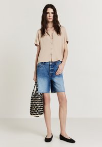 Blusa beige a maniche corte con bottoni, pantaloni corte in denim blu con orlo sfrangiato, borsa nera intrecciata e ballerine nere. Outfit semplice e casual.