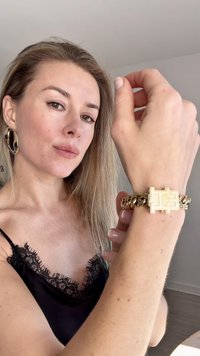 Goldenes Metallarmband mit geometrischem Verschluss, verwobenen Kettengliedern und glatter Oberfläche, getragen an einem Handgelenk mit einem schwarzen Spitzentop.