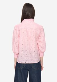 Lichtroze blouse met pofmouwen, oogletstof en een gerimpelde kraag. Heeft bloemmotieven met openingen en een losse pasvorm. Draagt bij donkere jeans.