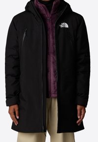 Abrigo negro aislante con capucha, que presenta un logotipo blanco y bolsillos con cremallera, superpuesto a una chaqueta de forro polar morado. Son visibles unos pantalones beige.