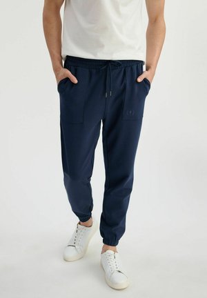 Mężczyzna ubrany w granatowe spodnie dresowe z troczkiem w talii i elastycznymi mankietami, w białych trampkach i białej koszuli, z rękami w kieszeniach.