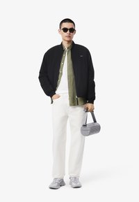 Zwarte bomberjack, olijfgroene shirt, wit T-shirt, crèmekleurige broek, grijze gewatteerde tas en grijze sneakers in een minimalistische setting.