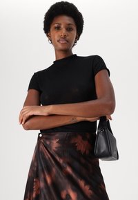 Selvsikker kvinne iført svart kortermet topp og brun blomstret skirt, holder en svart håndveske under armen mot en ensfarget hvit bakgrunn.