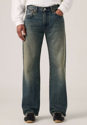 Mand iført falmede, løstsiddende blå jeans med forlommer, kombineret med sorte sneakers og en hvid langærmet top, hånd i lommen.