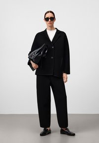 Blazer noir texturé avec boutons, chemise blanche, pantalon noir large et sac noir, associé à des chaussures plates noires et des lunettes de soleil.