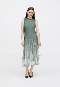 Vestido plissado sem mangas em verde, esbatendo para o branco. Listras verticais, tecido macio, comprimento até à canela, combinado com uma pequena mala branca e sapatos de salto pretos.