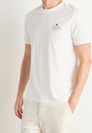 Mężczyzna w białej, krótkiej koszulce Tommy Hilfiger z logo na piersi, w połączeniu z jasnymi spodniami, stojący z jedną ręką w kieszeni.