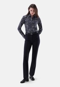 Camicia a maniche lunghe blu navy con un motivo paisley, abbinata a pantaloni neri su misura e stivaletti neri alla caviglia. Presenta un design aderente con colletto.