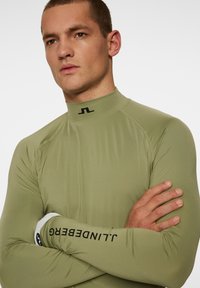 Långärmad sporttröja i olivgrön, med hög krage och svart logotyp vid halslinningen. Tyget har en slät, stretchig textur.