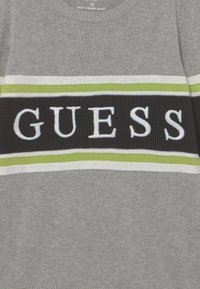 Harmaa collegepaita, jossa on leveä musta raita valkoisilla ja vihreillä yksityiskohdilla sekä sana "GUESS" valkoisilla kirjaimilla keskellä.