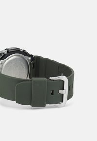 Digital watch with a black face, green textured silicone band, silver buckle, and stainless steel back. Features adjustable strap holes.