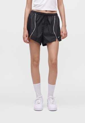 Vrouw draagt zwarte sportshorts met witte bies en een klein logo, witte sokken, witte sneakers en een wit cropped topje.