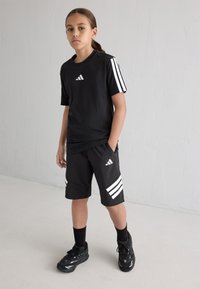 Μαύρη αθλητική μπλούζα με λευκό λογότυπο Adidas, συνδυασμένη με μαύρες βερμούδες με λευκές ρίγες και τσέπες, φορεμένη με μαύρα αθλητικά παπούτσια και κάλτσες.