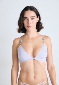 Vrouw met kort golvend bruin haar, gekleed in een licht paarse kanten bralette en bijpassende onderbroek tegen een effen lichte achtergrond.