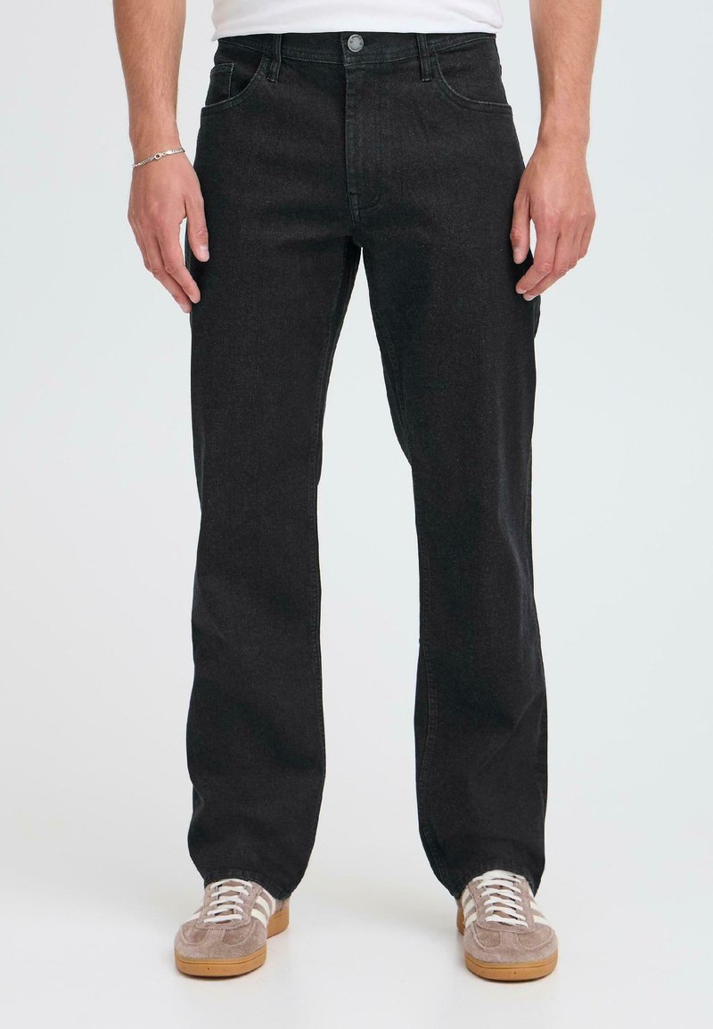 Jeans de mezclilla negra con un diseño de pierna recta, cinco bolsillos y cierre de botón, combinados con zapatillas grises que tienen cordones blancos.
