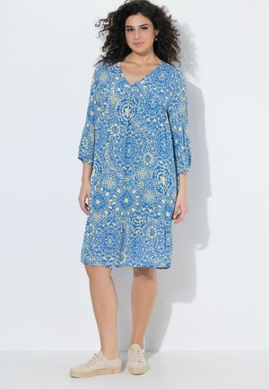 Kvinde med krøllet hår iført en blå og gul blomstermønstret knælange kjole og beige afslappede sko, stående mod en hvid baggrund.