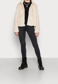 Chaqueta de piel sintética crema con capucha, suéter de punto negro, jeans negros ajustados y botas negras con cordones y suela gruesa.