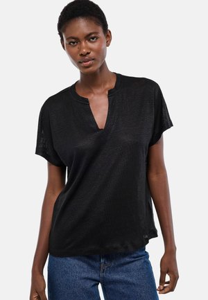 Femme portant un haut noir ample à manches courtes avec une encolure en V et un jean bleu, debout devant un fond clair uni.