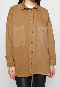 Giacca camicia oversize marrone in cotone con chiusura a bottoni, due tasche frontali e colletto. Tessuto liscio, design casual.