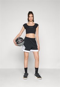 Svart kortärmad träningsöverdel med korta ärmar, i kombination med svarta shorts med vit kant och geometriskt mönster. En basketboll hålls i ena handen.