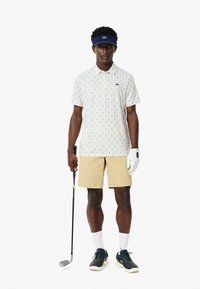 Valkoinen polo-paita, jossa on tummansinisiä kuvioita, yhdistettynä vaaleisiin beigeihin shortseihin. Asusteina on tummansininen visori, valkoinen käsine ja mustat golfkengät.