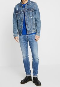Denim jacket with distressed details, button closure, and two chest pockets, worn over a blue t-shirt and paired with light wash jeans and black boots.