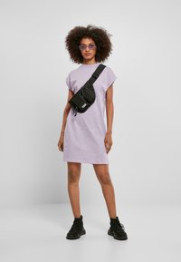 Robe lilas à manches courtes avec une coupe décontractée, associée à un sac bandoulière noir et des bottines noires. Le modèle a des cheveux bouclés et porte des lunettes de soleil.