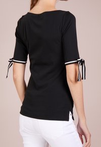 Černý krátký top s bílými pruhovanými akcenty na rukávech a bočních rozparcích. Vyrobený z hladkého materiálu s volným střihem. Zobrazen pohled zezadu.