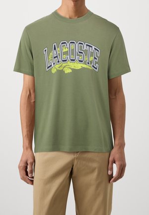 Mann trägt olivgrünes Lacoste-T-Shirt mit großem Logo und beiger Hose, dargestellt von Hals bis Mitte Oberschenkel vor neutralem Hintergrund.