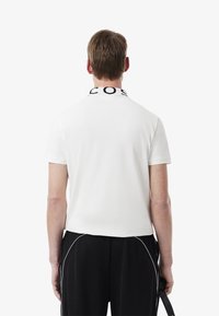 Vit kortärmad polo med krage och svart "COS"-logotyp, i kombination med svarta byxor med vit sidokant.