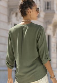 Olivgrüne Bluse mit Dreiviertelärmeln, ausgestattet mit einer Falte im Rücken und geknöpften Ärmeln. Glatte Textur, lockere Passform.