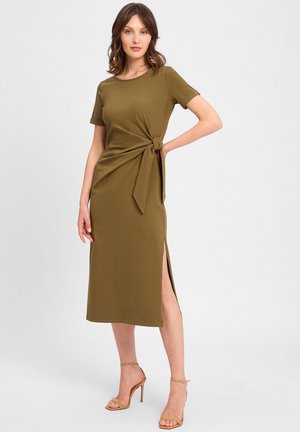Olivgrünes, kurzärmliges Kleid mit einem gebundenen Taillendetail, seitlichem Schlitz und glattem Stoff. Ergänzt durch beigefarbene, hochhackige Sandalen.