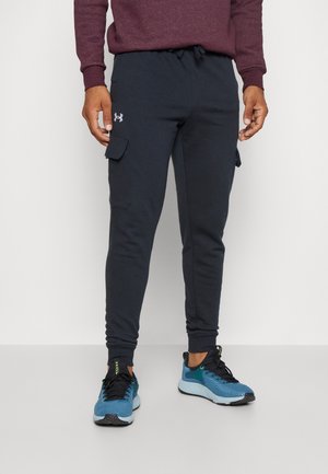 Bas du corps d'une personne portant un pantalon cargo jogger noir avec le logo Under Armour et des chaussures de course bleues, sur un fond clair uni.