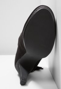 Bottine noire en daim avec talon fin, semelle texturée et détail pailleté le long du bord, positionnée semelle vers le haut contre un mur blanc.