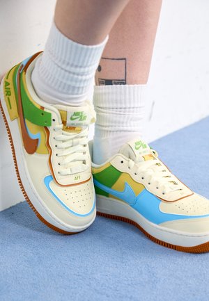 Beine tragen weiße Socken und bunte Nike Air Force 1 Sneakers mit grünen, blauen, gelben und braunen Akzenten auf blauem Boden nahe einer weißen Wand.
