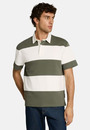 Homme portant un polo manches courtes à larges rayures vertes et blanches et un jean bleu, regardant vers sa droite.